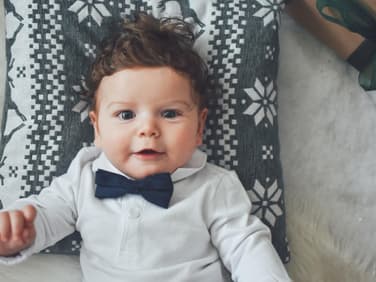 Kleiner Junge mit Fliege lächelt auf einem Kissen liegend in die Kamera.