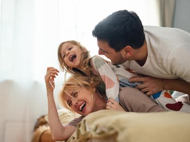 Eltern und Kind liegen im Familienbett und lachen.
