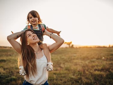 Mutter trägt Kind auf einer Wiese auf den Schultern.