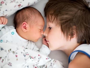 Großer Bruder liegt neben Baby im Bett.