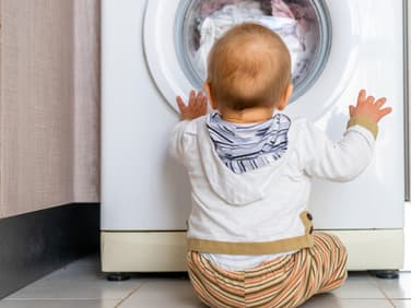 Baby sitzt vor der Waschmaschine und schaut hinein.